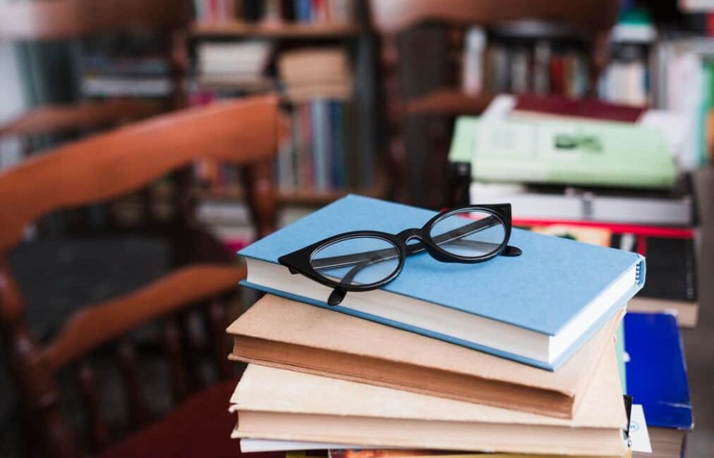 A pair of glasses atop a blue book on a pile, with a blurred bookshelf behind