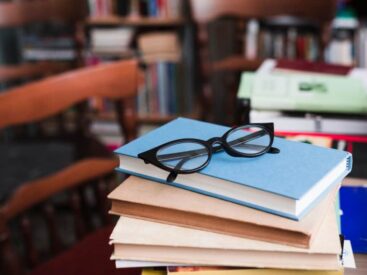 A pair of glasses atop a blue book on a pile, with a blurred bookshelf behind