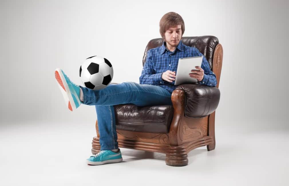 A man casually balances a soccer ball on his foot while using a tablet