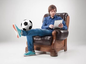 A man casually balances a soccer ball on his foot while using a tablet