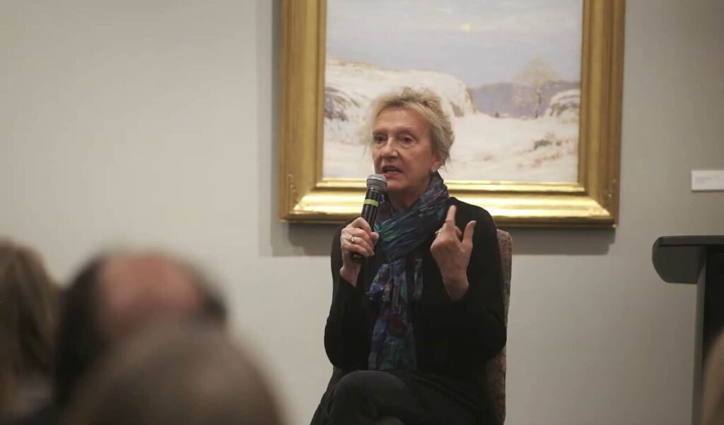 A woman speaking into a microphone with a painting behind her